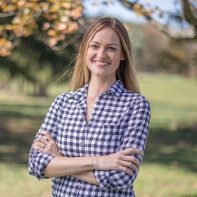 Square head shot of Paige Gebhardt dressed in a plaid shirt, standing in an orchard