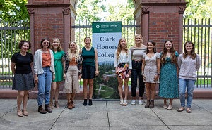 A group of students standing in front of a wrought iron fence of an outdoor courtyard in front of a sign reading "Clark Honors College." 