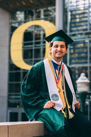 Undergraduate in green graduation cap and gown, sitting outside with an Oregon O in the background.
