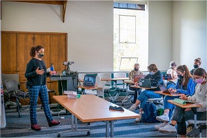 An instructor, masked against COVID, presents a class to masked students and a laptop's web-camera.