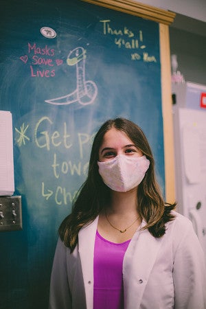 A young woman with long hair, in a floral-print COVID mask, wearing a white jacket over a fuchsia top, stands in front of a blackboard.