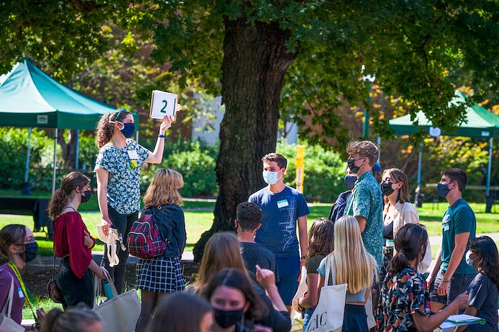 A group of students, masked against COVID, gather around a guide on a sunny day under an oak.