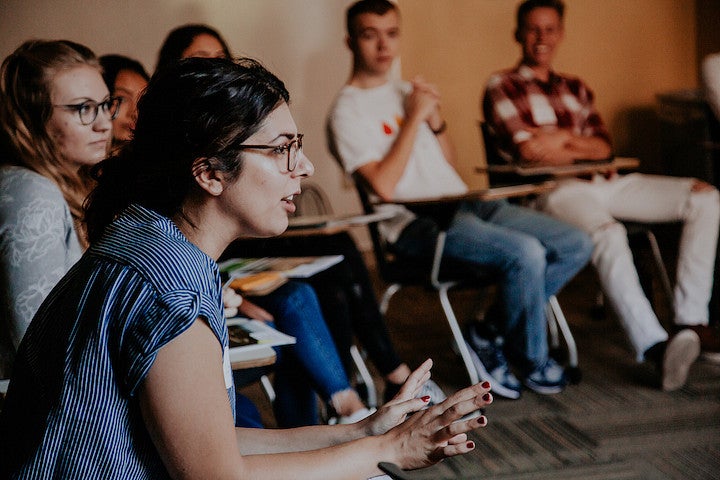 A woman leans forward in her chair while speaking to a circle of seated students.