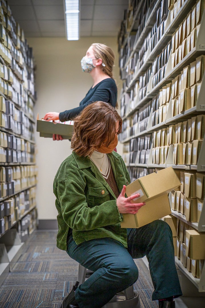Two women, masked against COVID, research materials between two library shelves.