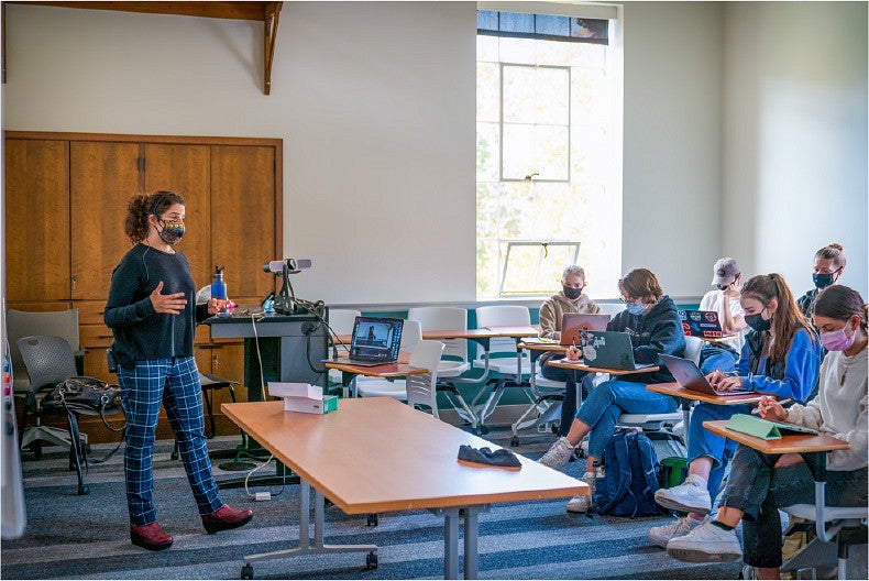 An instructor, masked against COVID, presents a class to masked students and a laptop's web-camera.