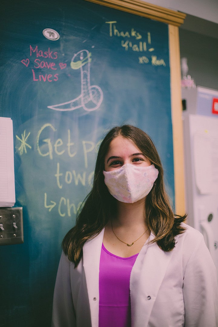 A young woman with long hair, in a floral-print COVID mask, wearing a white jacket over a fuchsia top, stands in front of a blackboard.