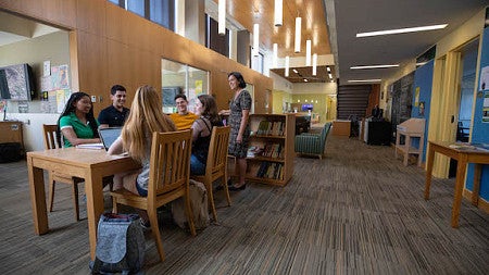 Long hallway with students with laptops seated around a table; one person is standing.