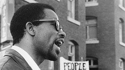 a black and white profile of a black man speaking in front of brick buildings.
