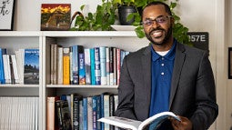 Marc Robinson smiling and standing in front of a book shelf while holding a book.