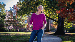 Lindsay Hinkle in a pink sweater standing under U of O campus oaks turning red in early autumn.