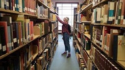 A student standing between library shelves reaches for a book.