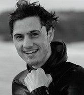 Black and white head shot of Aaron Georis in a wetsuit, blurred body of water in background.